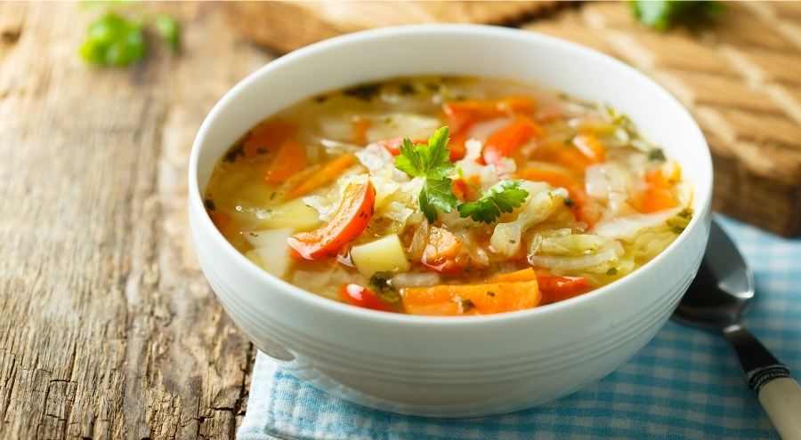 A vertical, top-down shot of a vibrant, chunky cabbage soup in a rustic bowl with a gold spoon, surrounded by fresh herbs like parsley and celery