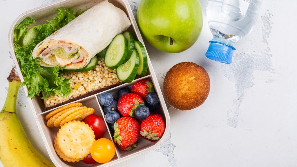 Balanced packed lunch with a wrap, cucumber slices, grains, crackers, berries, banana, apple, muffin, and water bottle.