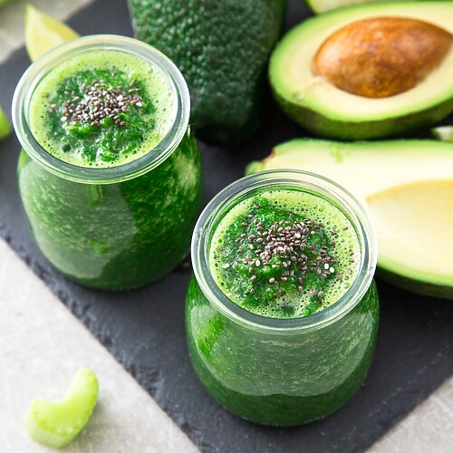 Green avocado smoothie with chia seeds, served in glass jars beside avocado and lime slices.