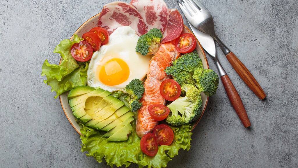 High-protein salad bowl with salmon, egg, avocado, broccoli, tomatoes, lettuce, and sliced meat on a gray table.