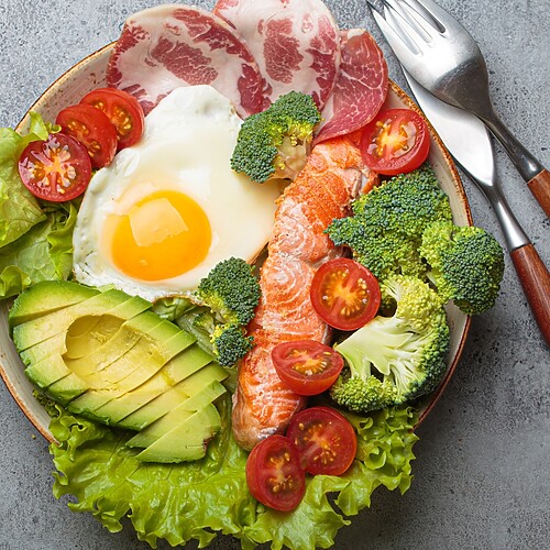 High-protein salad bowl with salmon, egg, avocado, broccoli, tomatoes, lettuce, and sliced meat on a gray table.