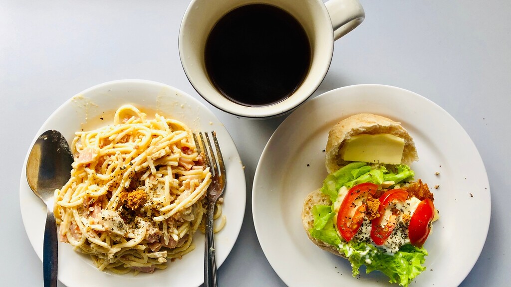 Overhead view of spaghetti, an open sandwich with lettuce and tomato, and a cup of black coffee on white plates.