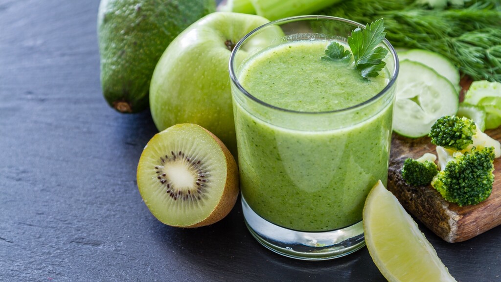 Green smoothie with kiwi, avocado, cucumber, broccoli, lime, and fresh herbs on a dark surface.