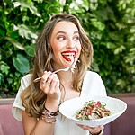 Woman eating a fresh salad outdoors with leafy green plants in the background.