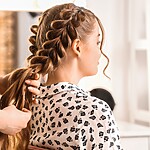 Hairstylist braiding long brown hair into a loose Dutch braid on a woman seated in a salon.