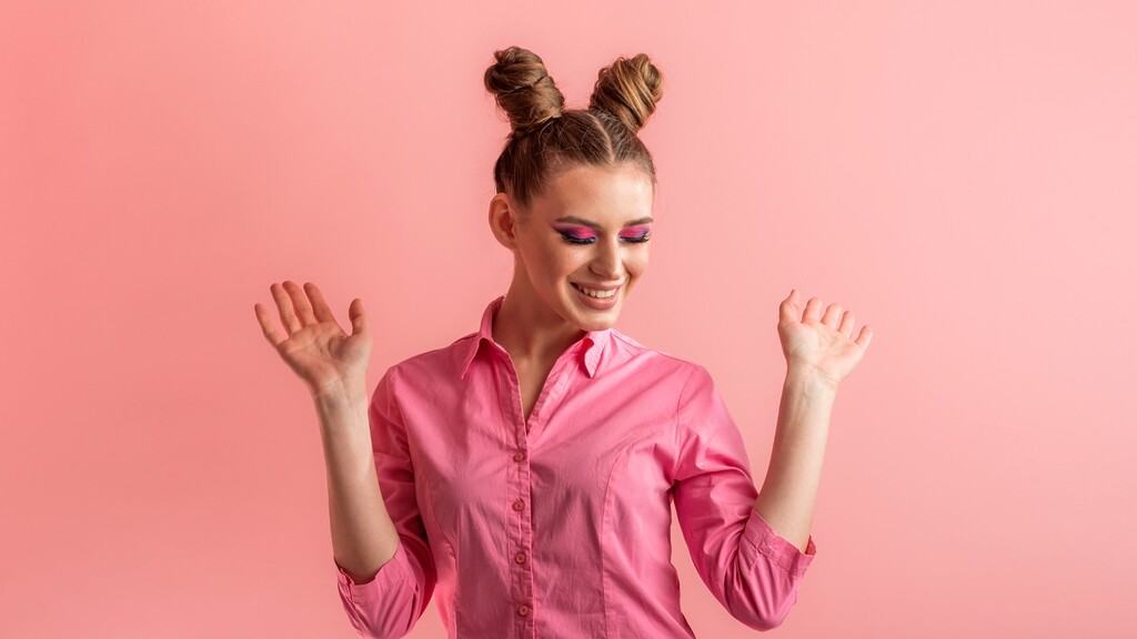 Smiling woman with double space buns hairstyle wearing a pink shirt against a pink studio background.