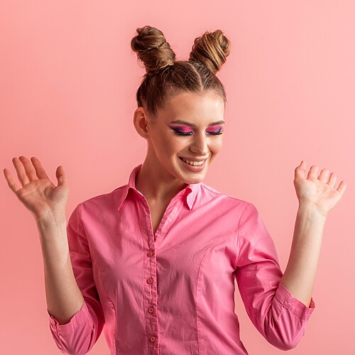 Smiling woman with double space buns hairstyle wearing a pink shirt against a pink studio background.