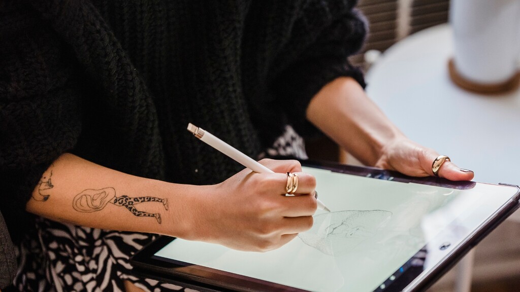Person with forearm tattoos sketching a custom tattoo design on a digital tablet with a stylus.