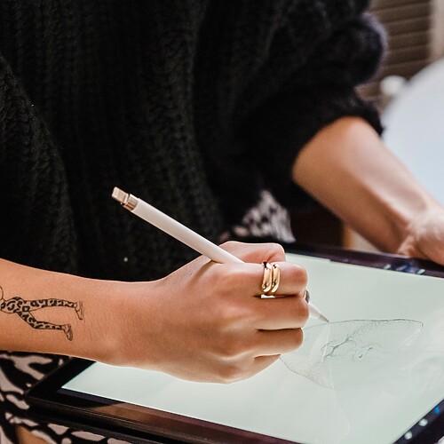 Person with forearm tattoos sketching a custom tattoo design on a digital tablet with a stylus.