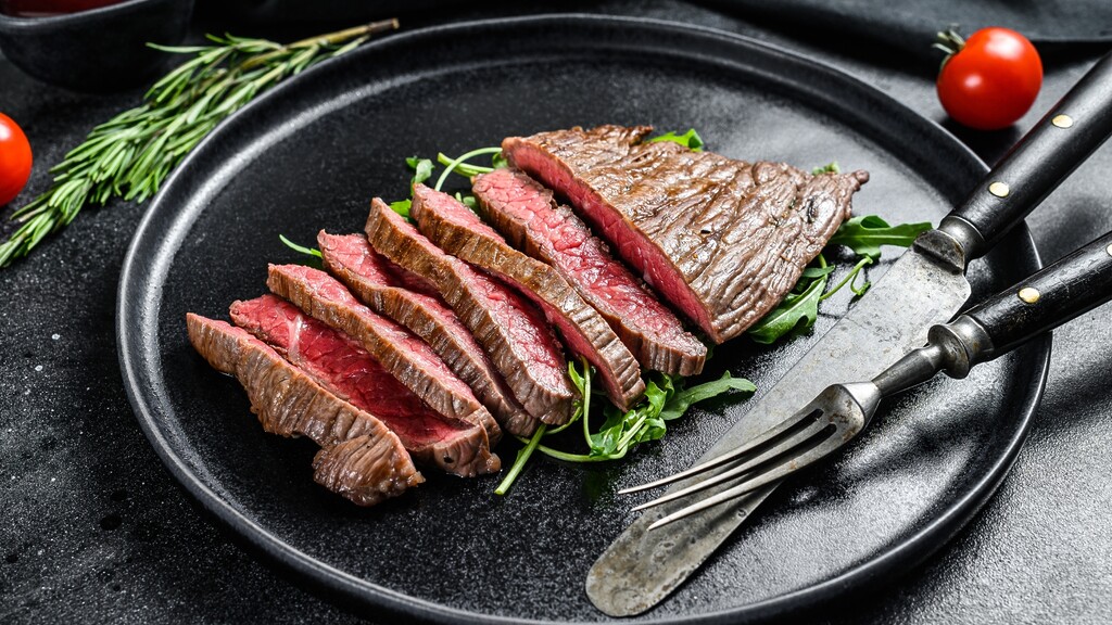 Sliced lean steak served on a black plate with greens and utensils.