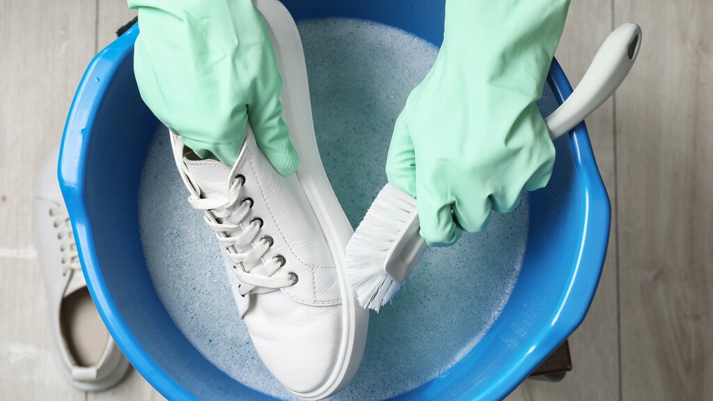 Hands scrubbing a white sneaker with a brush over soapy water.