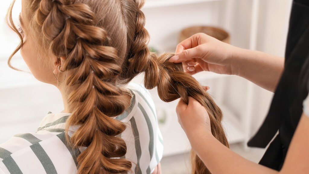 Hairstylist creating a voluminous bubble braid on long auburn hair, showing the elastic sections being shaped by hand.