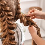 Hairstylist creating a voluminous bubble braid on long auburn hair, showing the elastic sections being shaped by hand.