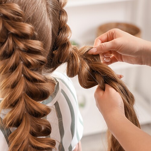 Hairstylist creating a voluminous bubble braid on long auburn hair, showing the elastic sections being shaped by hand.