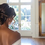 22 Easy Bun Hairstyles for Every Occasion and Hair Type Woman in a white off-shoulder dress showing a romantic low curly bun hairstyle in a bright dressing room.