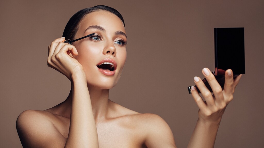 Woman applying mascara while looking into a compact mirror during her makeup routine.