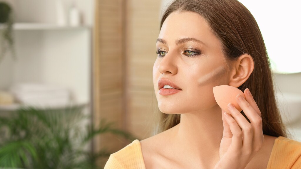 Woman applying cream contour with a makeup sponge to softly sculpt her cheekbones for a natural date night look.