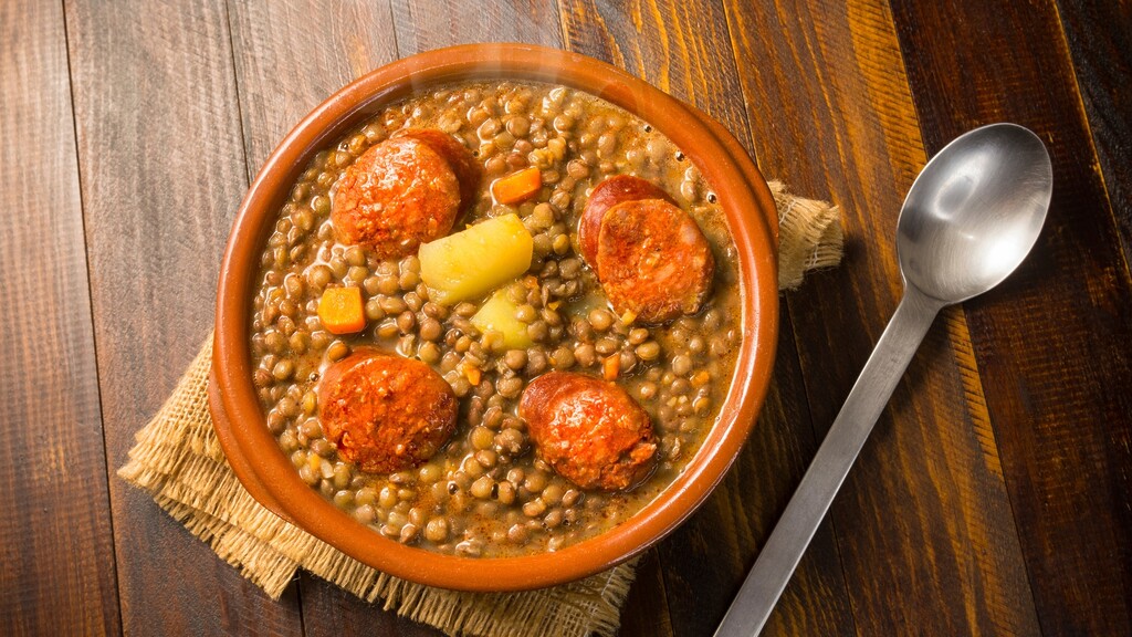 Bowl of cooked lentils with vegetables and sausage on a wooden table.