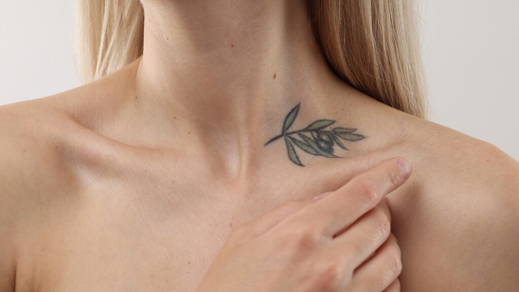 Alt text: Close-up of a woman pointing to a delicate olive branch tattoo near her collarbone.