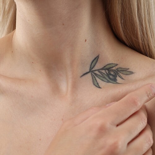 Alt text: Close-up of a woman pointing to a delicate olive branch tattoo near her collarbone.