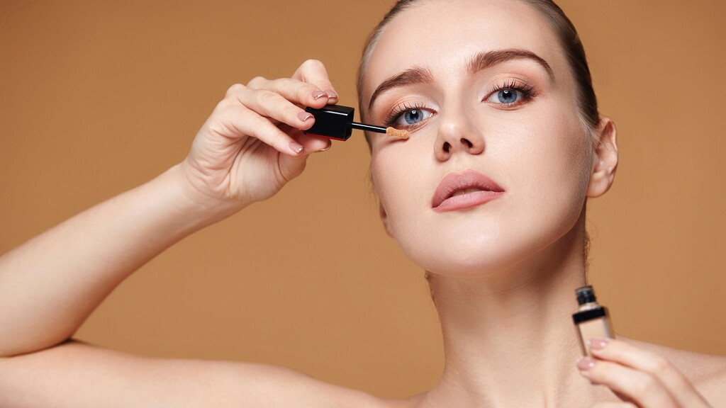 Woman applying liquid concealer under her eye with a makeup wand against a warm neutral background.