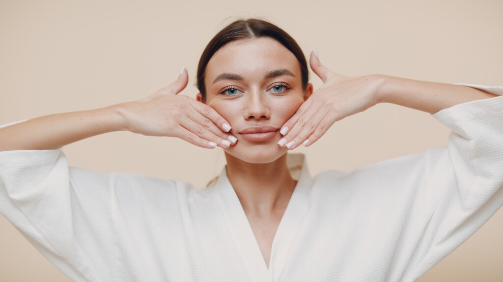 Young woman in a white robe demonstrating a face yoga cheek lift massage, with both hands placed along her jawline and cheeks against a neutral background.