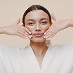 Young woman in a white robe demonstrating a face yoga cheek lift massage, with both hands placed along her jawline and cheeks against a neutral background.