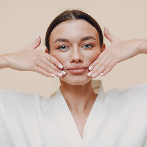 Young woman in a white robe demonstrating a face yoga cheek lift massage, with both hands placed along her jawline and cheeks against a neutral background.