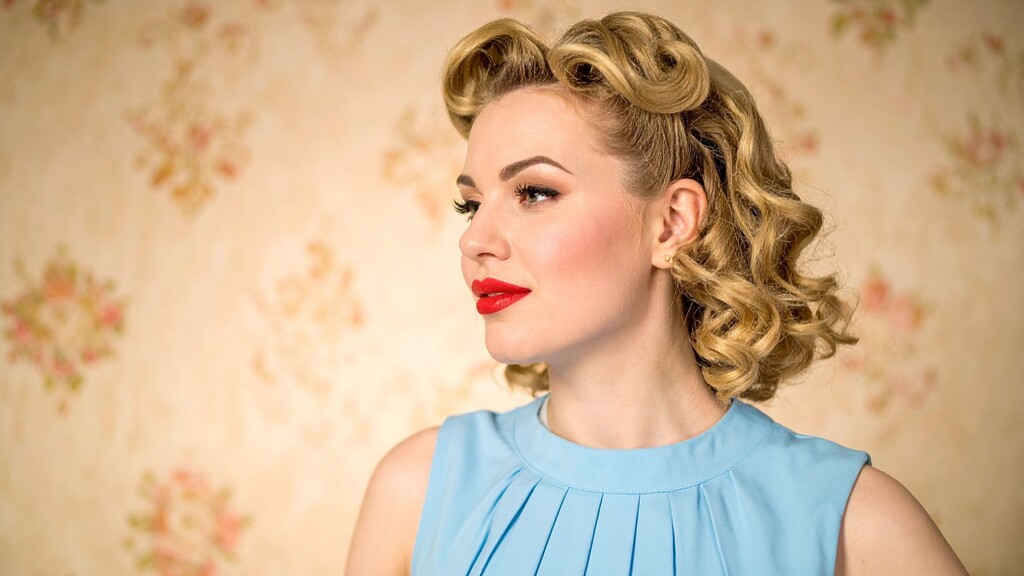 Woman with blonde vintage curls and red lipstick wearing a blue dress against a soft floral background.