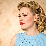 Vintage Hairstyles for Different Hair Lengths (That Actually Hold) Woman with blonde vintage curls and red lipstick wearing a blue dress against a soft floral background.