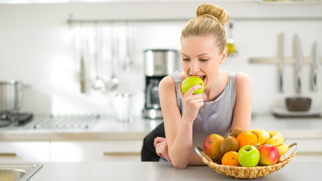 Woman eating a green apple in a bright kitchen beside a basket of fresh fruit.