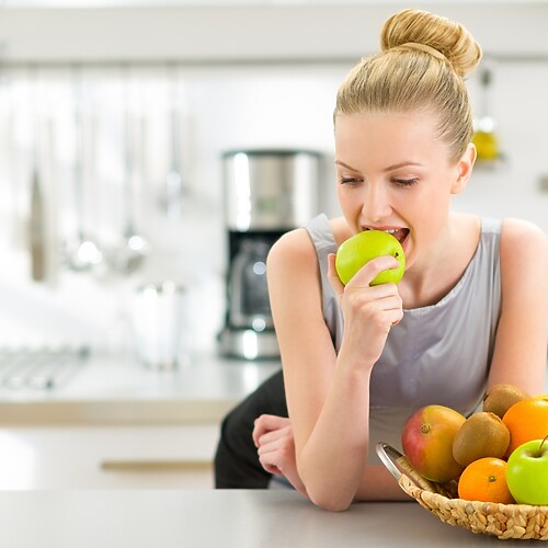 Woman eating a green apple in a bright kitchen beside a basket of fresh fruit.