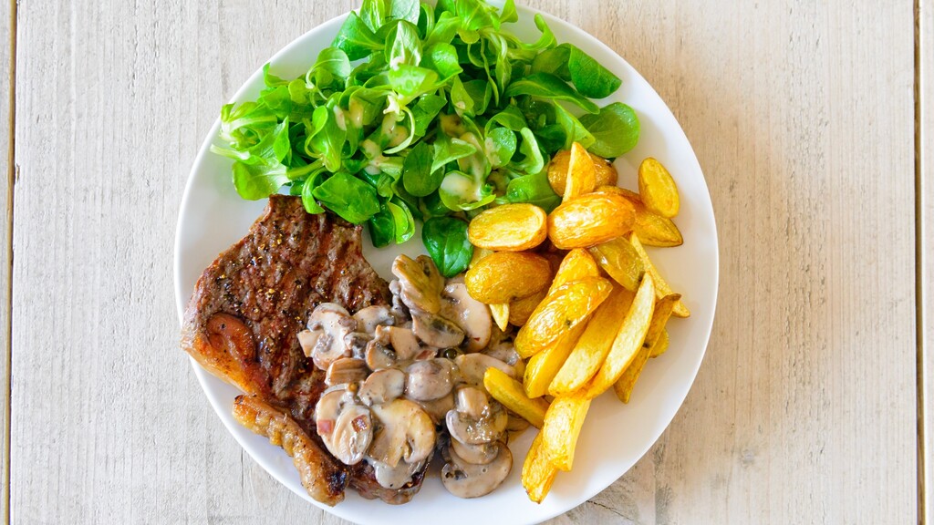 Balanced dinner plate with steak, leafy green salad, roasted potatoes, and mushrooms on a wooden table.