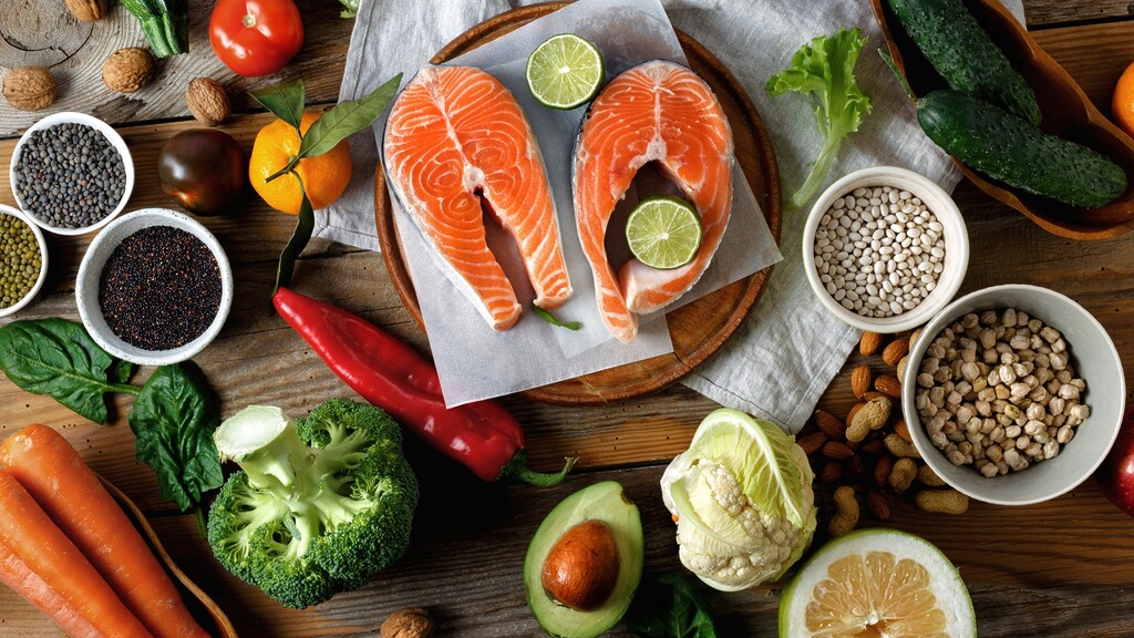 Fresh hormone balancing foods including salmon, avocado, broccoli, beans, lentils, nuts, and leafy greens on a wooden table.