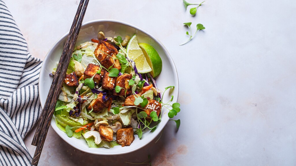 Tofu salad bowl with leafy greens, lime wedges, sesame seeds, and chopsticks.