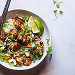 Tofu salad bowl with leafy greens, lime wedges, sesame seeds, and chopsticks.