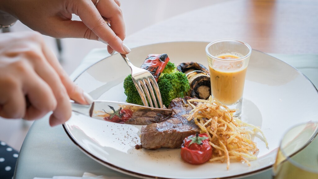 A person cutting a meal with steak, broccoli, roasted vegetables, and sauce on a white plate.