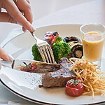 A person cutting a meal with steak, broccoli, roasted vegetables, and sauce on a white plate.