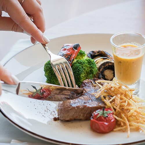 A person cutting a meal with steak, broccoli, roasted vegetables, and sauce on a white plate.