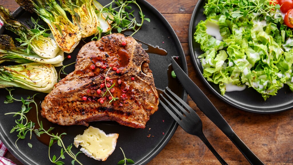 Grilled pork chop with roasted bok choy, herbs, soft cheese, and a side green salad on a wooden table.