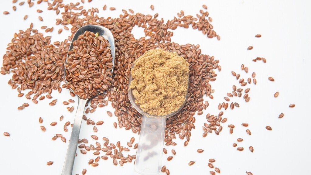 Whole and ground flaxseeds in measuring spoons on a white surface.
