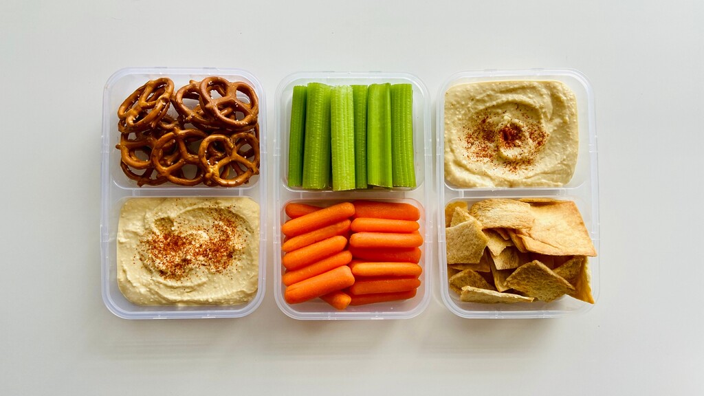Meal prep containers with hummus, pretzels, celery sticks, baby carrots, and pita chips arranged on a white table.