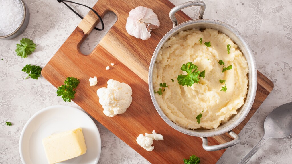Creamy cauliflower mash with garlic, butter, parsley, and cauliflower florets on a cutting board.