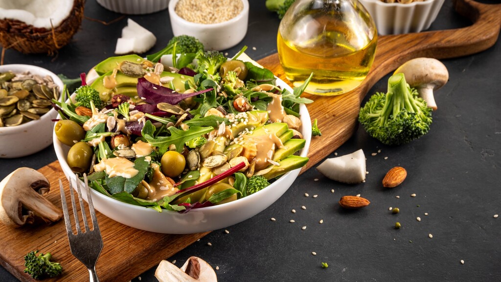 Keto-friendly green salad with avocado, broccoli, olives, seeds, almonds, mushrooms, coconut, and olive oil on a dark surface.