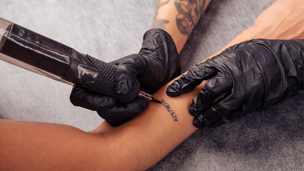 Tattoo artist wearing black gloves creating a small text tattoo on a client’s arm with a tattoo machine