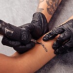 Tattoo artist wearing black gloves creating a small text tattoo on a client’s arm with a tattoo machine