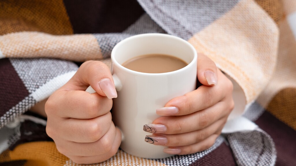 Hands holding a warm mug of coffee under a cozy plaid blanket.