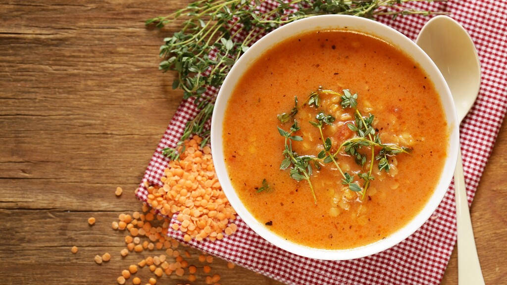 Lentil soup in a white bowl with herbs, red lentils, and a spoon on a wooden table.
