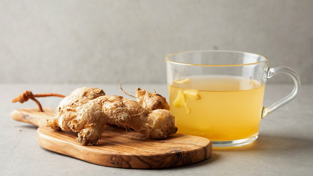 Fresh ginger root beside a clear cup of ginger tea on a small wooden board.