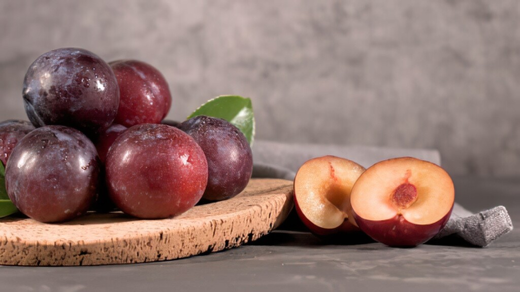Fresh plums on a wooden board, with one plum sliced in half beside a gray cloth.